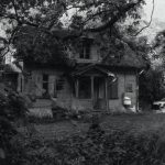 Eerie black and white photo of an abandoned house surrounded by dense foliage and trees.
