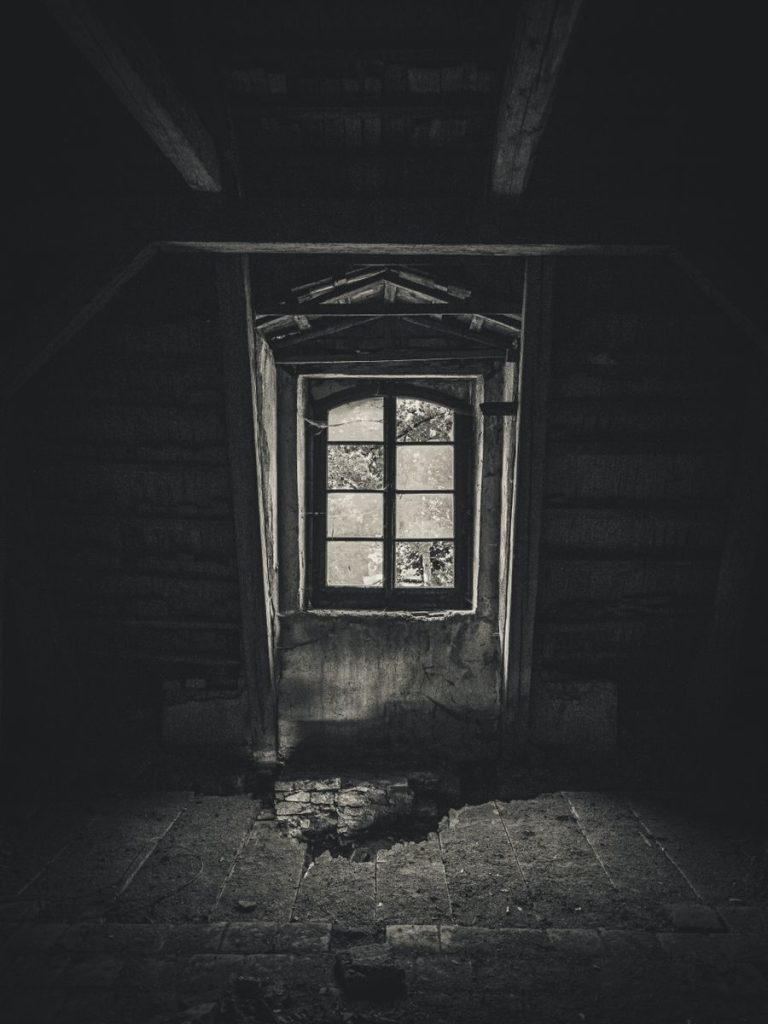 A haunting monochrome image of an abandoned attic, featuring a broken window and eerie shadows.
