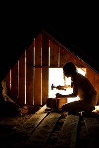 Silhouette of a woman hammering in a wooden attic workshop, backlit by warm light.