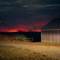 Peaceful countryside scene with a dramatic sunset over a farmhouse and gravel road.