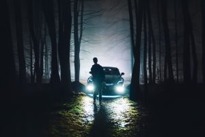 A silhouette of a man standing in front of a car with headlights on in a foggy forest at night.