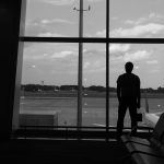 A silhouette of a man standing at an airport terminal window, looking onto the tarmac.