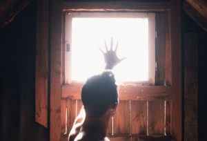 A dramatic silhouette of a man reaching towards a bright window with wooden surroundings.
